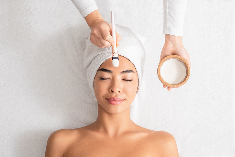Beauty Treatments. Cosmetologist applying clay mask on asian woman face at spa salon